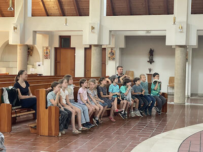 Eine Studentin und ein Schulassistent sitzen mit den Kindern in den Kirchenbänken und hören den Erzählungen der Religionslehrerin zu.