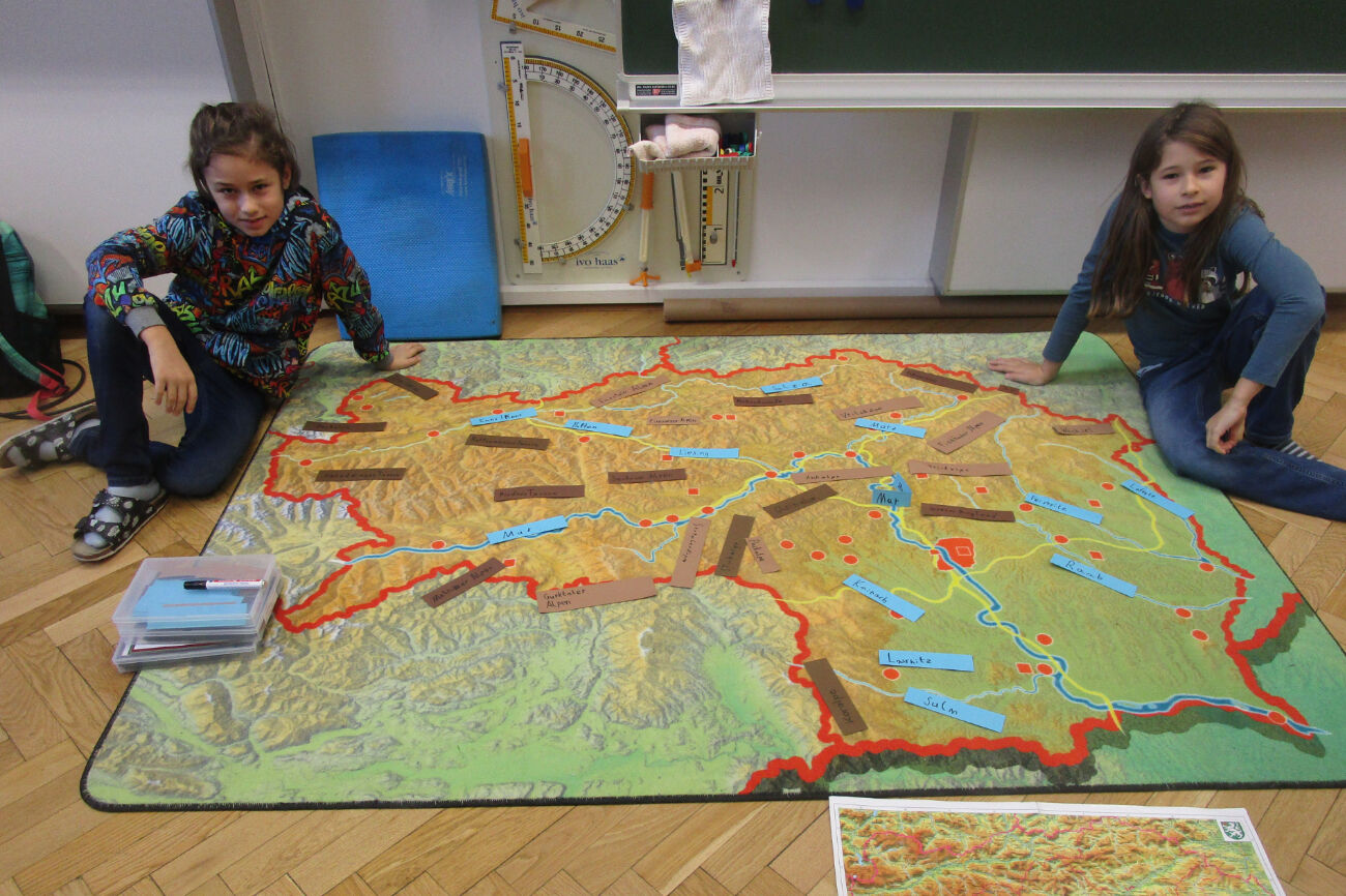 Auf einem Steiermark-Teppich haben zwei Buben wichtige Flüsse und Gebirge mit Kärtchen markiert.